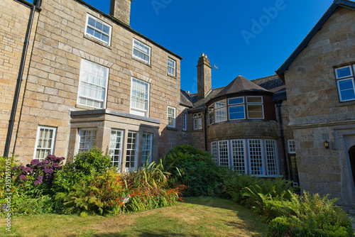 A historic, multi-story stone building features a prominent bay window. Green gardens surround the structure under a clear blue sky - Kenegie Manor, Cornwall, UK
