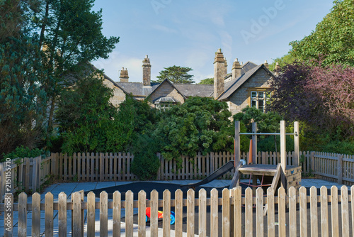A charming stone building peeks through dense greenery, overlooking a vibrant children's playground. A wooden fence encloses the play area - Kenegie Manor, Cornwall, UK