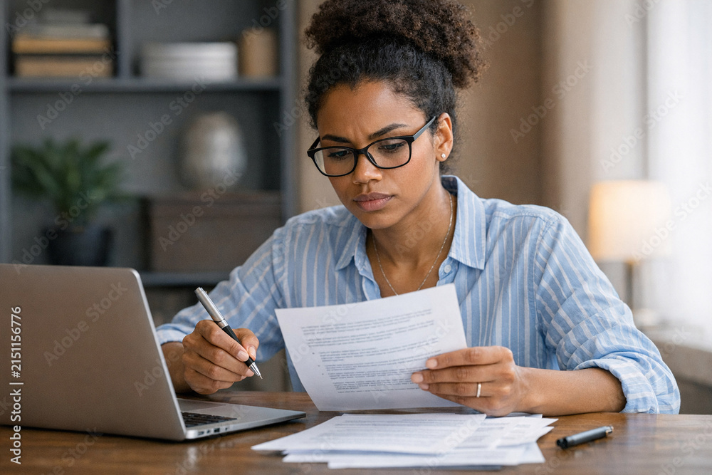 Fototapeta premium A professional woman in glasses working diligently on digital and printed documents at her desk, with a focus on efficiency and organization.