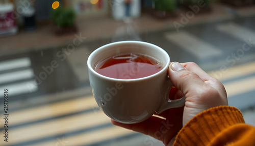 Hand holding a steaming mug of hot tea by a window with a blurred street view, symbolizing comfort and warmth.