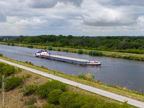 Cargo barge travels along Mittellandkanal through green landscape beneath dramatic summer clouds. Inland waterway supports freight transport, navigation and logistics across northern Germany
