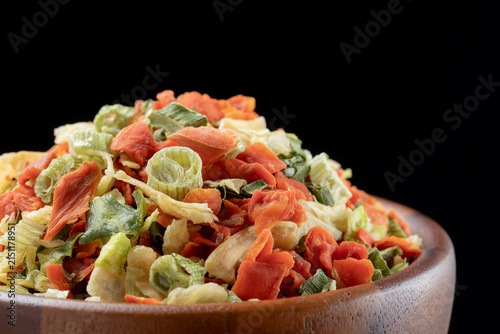 Pile of dehydrated shredded vegetables  (cabbage, green onions, and carrots) in a brown wooden bowl on black background.