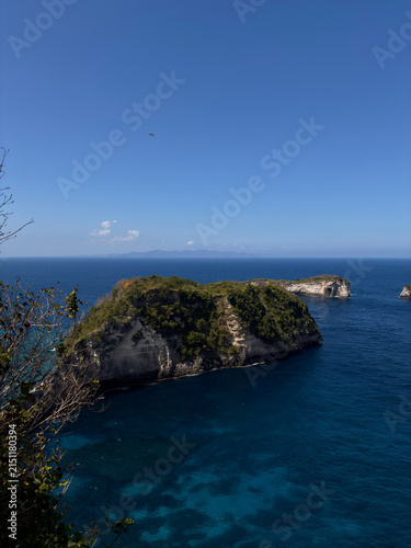 A scenic view of rocky islands in a clear blue ocean. The coastline features lush green vegetation contrasting with the vibrant water. The sky above is bright and sunny.