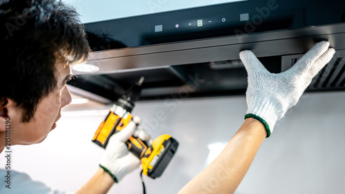 Asian male technician using electric screwdriver fixing extractor motor in cooker hood in modern kitchen. Maintenance worker man repairing exhaust fan in kitchen hood. Home appliance service