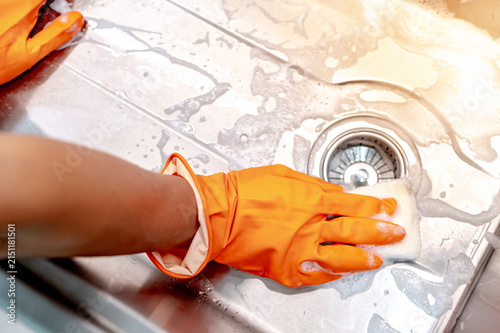 Obraz na plátně Housekeeper hand wearing orange rubber glove scrubbing on kitchen sink drainer with soap suds spreading around