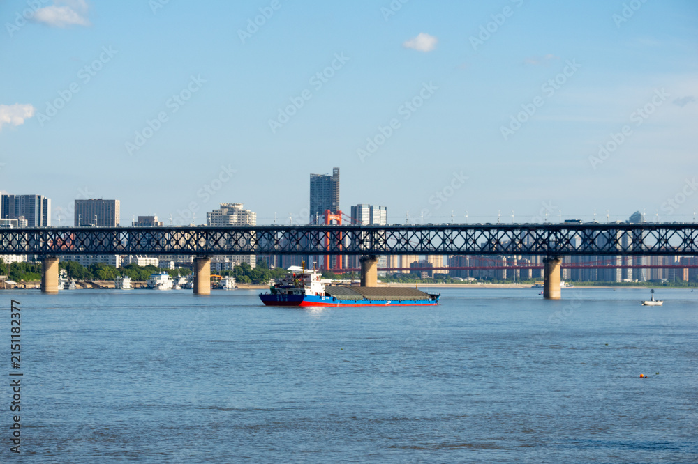 Obraz premium A cargo ship sails on the Yangtze River under the large steel bridge in the city of Wuhan China during a bright sunny day