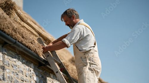 A thatcher works a steep cottage roof under harsh midday sun, bundles of fresh reed stacked along the eaves as a Valentines Day red rose bouquet sits propped against the ladder in