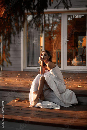 A Caucasian woman, wrapped in a blanket, sits on the porch of a cozy house in autumn. Sunset in the autumn forest, outdoors. Romantic.