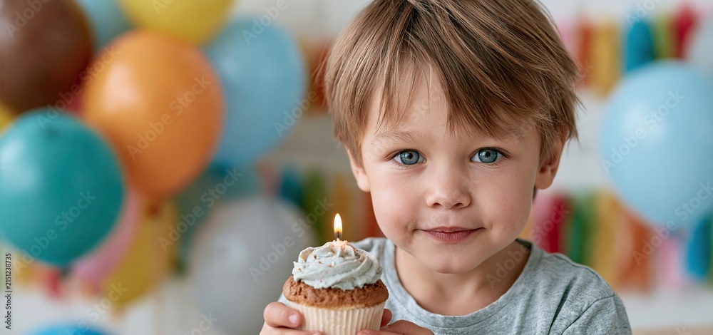 Obraz premium Toddler enjoying a birthday cupcake with lit candle surrounded by colorful balloons and festive decorations.