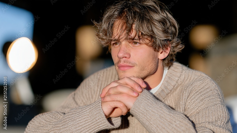 Obraz premium Concerned young man in a beige knit sweater sitting indoors with clasped hands, ideal for anxiety, decision making and mental health concepts.