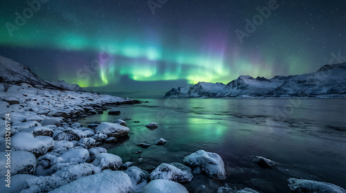 Breathtaking northern lights dancing above snowy mountain peaks reflecting in icy arctic ocean water on clear winter night.