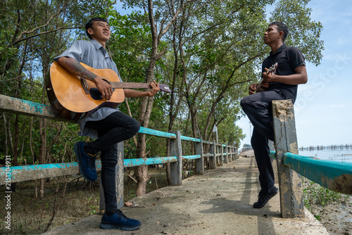Two friends leaning on bridge railing playing acoustic guitar and ukulele. Two musicians enjoying playing music together on a coastal bridge.