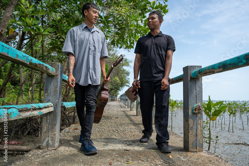Asian friends carrying guitar and ukulele walking on coastal bridge. Two musicians strolling along a bridge near a mangrove forest while holding their instruments on a sunny day.