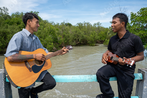 Two friends leaning on bridge railing playing acoustic guitar and ukulele. Two musicians enjoying playing music together on a coastal bridge.