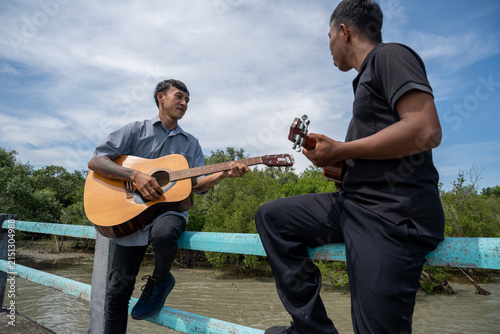 Two friends leaning on bridge railing playing acoustic guitar and ukulele. Two musicians enjoying playing music together on a coastal bridge.