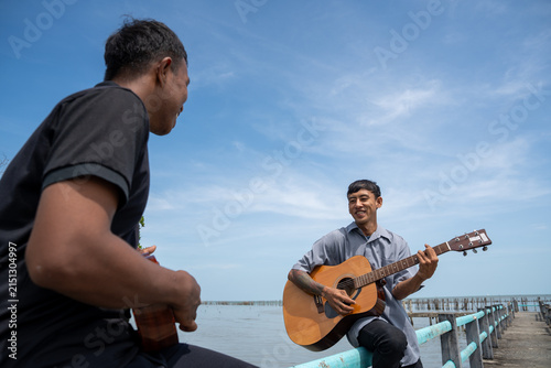 Two friends leaning on bridge railing playing acoustic guitar and ukulele. Two musicians enjoying playing music together on a coastal bridge.