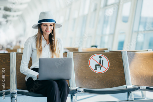 Woman wearing hat works on laptop computer while sitting in airport waiting area with social distancing sign on seat
