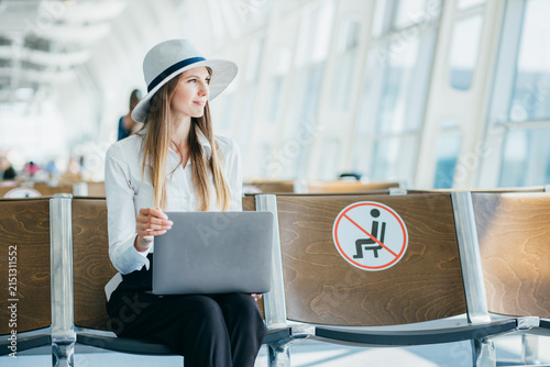 Woman wearing hat and white shirt sits with laptop in airport waiting area near social distancing sign