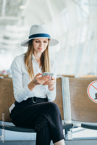 Young woman wearing hat and white shirt sits in airport waiting area using smartphone while waiting for flight
