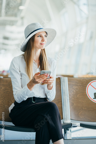 Woman wearing hat and white shirt holding smartphone while sitting in airport waiting area