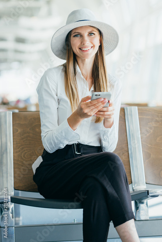 Young woman wearing hat and white shirt sits in airport lounge holding smartphone and smiling