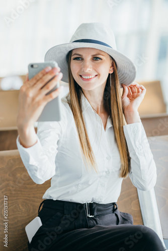 Young woman wearing hat and white shirt takes selfie with smartphone while sitting indoors