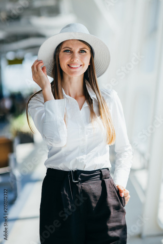 Young woman wearing wide-brimmed hat and white button-up shirt with dark trousers, smiling confidently