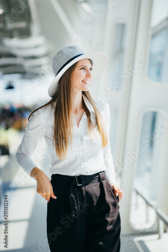 Young woman wearing a white shirt and black pants with a hat stands near a window looking outside