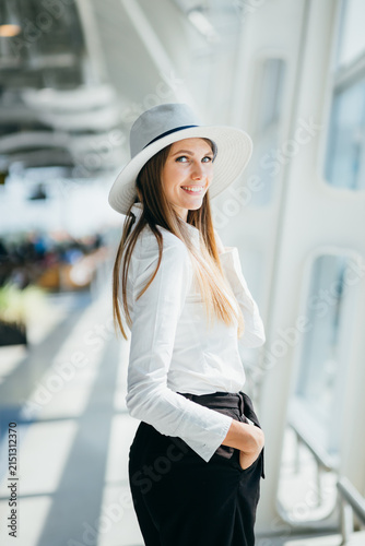 Young woman wearing wide-brimmed hat and white shirt stands with hand in pocket inside modern building