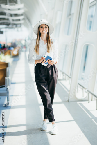 Smiling woman in hat and white shirt holding passport and boarding pass in airport terminal
