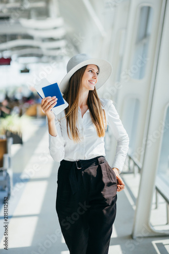 Smiling woman wearing a hat and elegant clothes holds passport and ticket in airport terminal