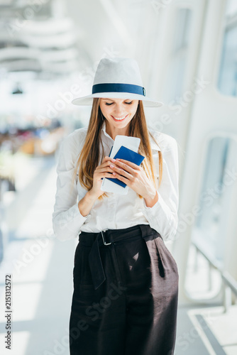 Smiling woman wearing a white hat and shirt holds passport and ticket while standing in bright airport terminal
