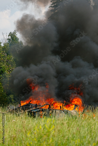 Vertical shot of a burning car wreck in a green grassy field with bright orange flames and huge black smoke column rising above trees on a summer day