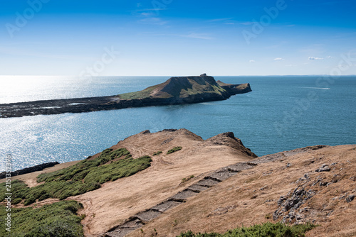 A high viewpoint overlooks Worm's Head and the calm summer sea, with drought-dried grassland in the foreground and sunlight sparkling across the water.