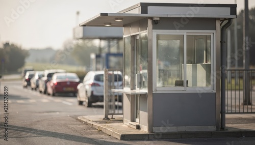 Daylight medium shot of a ticket booth at the entrance gate with clear focus while vehicles and surroundings softly fade out in a muted matinee glow.