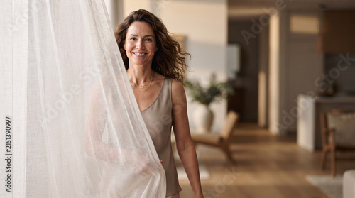 A smiling middle aged woman standing by a bright window at home.