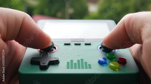 Close-up of female gamer hands holding and playing a green retro portable video game console outdoors against a blurred nature background.