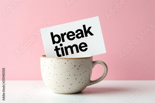 White paper note with break time text inside ceramic mug. Minimalist concept for rest period office pause. Pink background creates bright mood for morning routine relaxation during work day. Simple