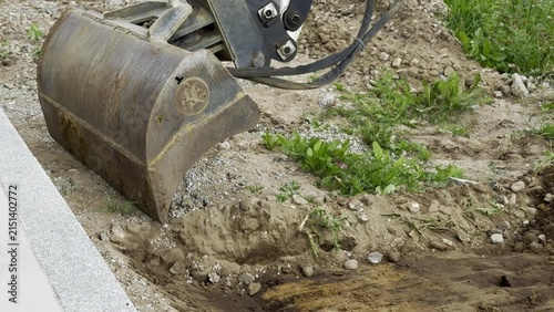 Excavator bucket hovers over a dirt mound at a construction site, with grass and gravel in the foreground 
