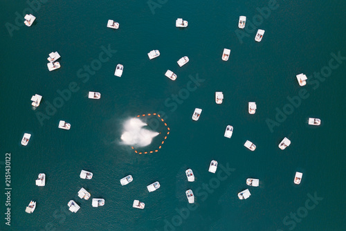 An aerial view of numerous small boats dispersed across deep blue-green water, surrounding a central boat that is producing a visible wake.