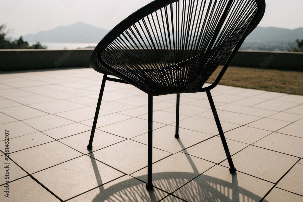 Obraz premium Modern black metal chair with string seat sits on tiled patio. Scenic view of mountains, sea in background. Outdoor furniture design for terrace garden relaxation. Minimalist patio decor with