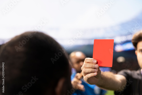Fotografie Football referee holds a red card while players protest the dismissal decision, creating controversy over unsportsmanlike conduct and a dramatic send-off during the soccer match on the stadium pitch