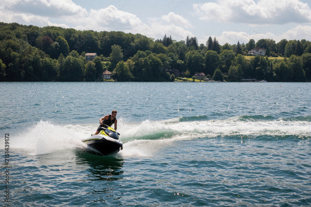 Fototapeta premium Young man rides fast jet ski on summer lake water. Male person makes sharp turn with motorboat creates big splash spray wave. Active water sport lifestyle on sunny day near green forest shore. Person