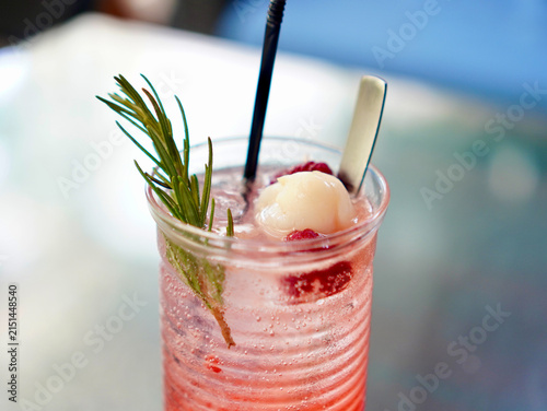 Close-up of cold sparkling lychee raspberry drink served in a clear glass with ice, spoon, and rosemary sprig
