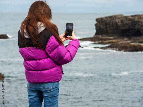Girl in pink jacket taking photo of rocky coastline. She stands near ocean with waves crashing. Her long hair flows in breeze. She wears blue jeans and holds smartphone. Seagulls fly above cliffs