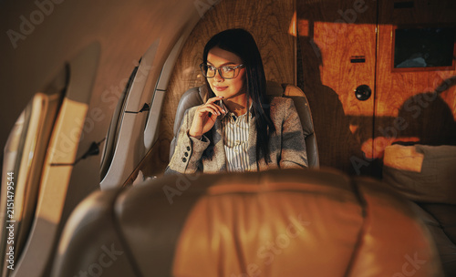 A dark haired woman, wearing a tweed blazer and striped shirt, sits with her hand raised to her chin in a leather seat of a private jet, surrounded by warm wood paneled walls bathed in amber light.