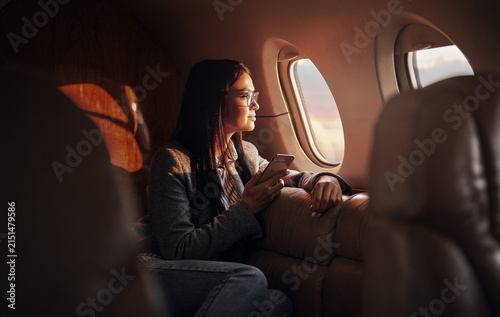 A dark haired woman, wearing glasses and a grey blazer, holds a smartphone and gazes quietly out the oval window of a private jet, her face lit by deep warm amber tones from outside.