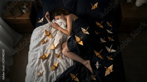 A peaceful young woman sleeping deeply in bed surrounded by shimmering golden origami paper cranes