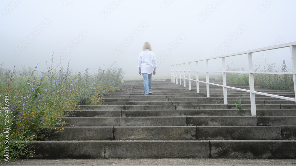 Obraz premium Woman walking up concrete stairs into dense fog. Solitary person in white shirt and jeans ascending outdoor steps towards the misty unknown