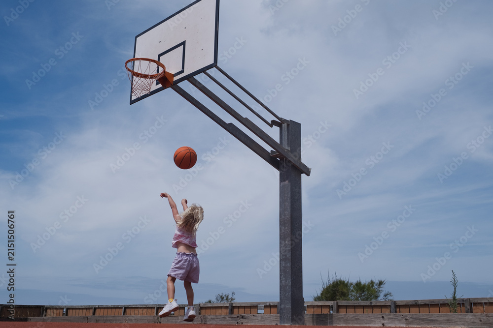 Fototapeta premium Blonde Toddler Girl Jumping to Shoot a Ball on an Outdoor Basketball Court